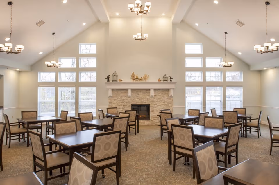 A spacious dining room with multiple dark wood tables and cushioned chairs arranged neatly. The room features a high vaulted ceiling with several hanging light fixtures, large windows letting in natural light, and a stone fireplace with decorative items on the mantel.