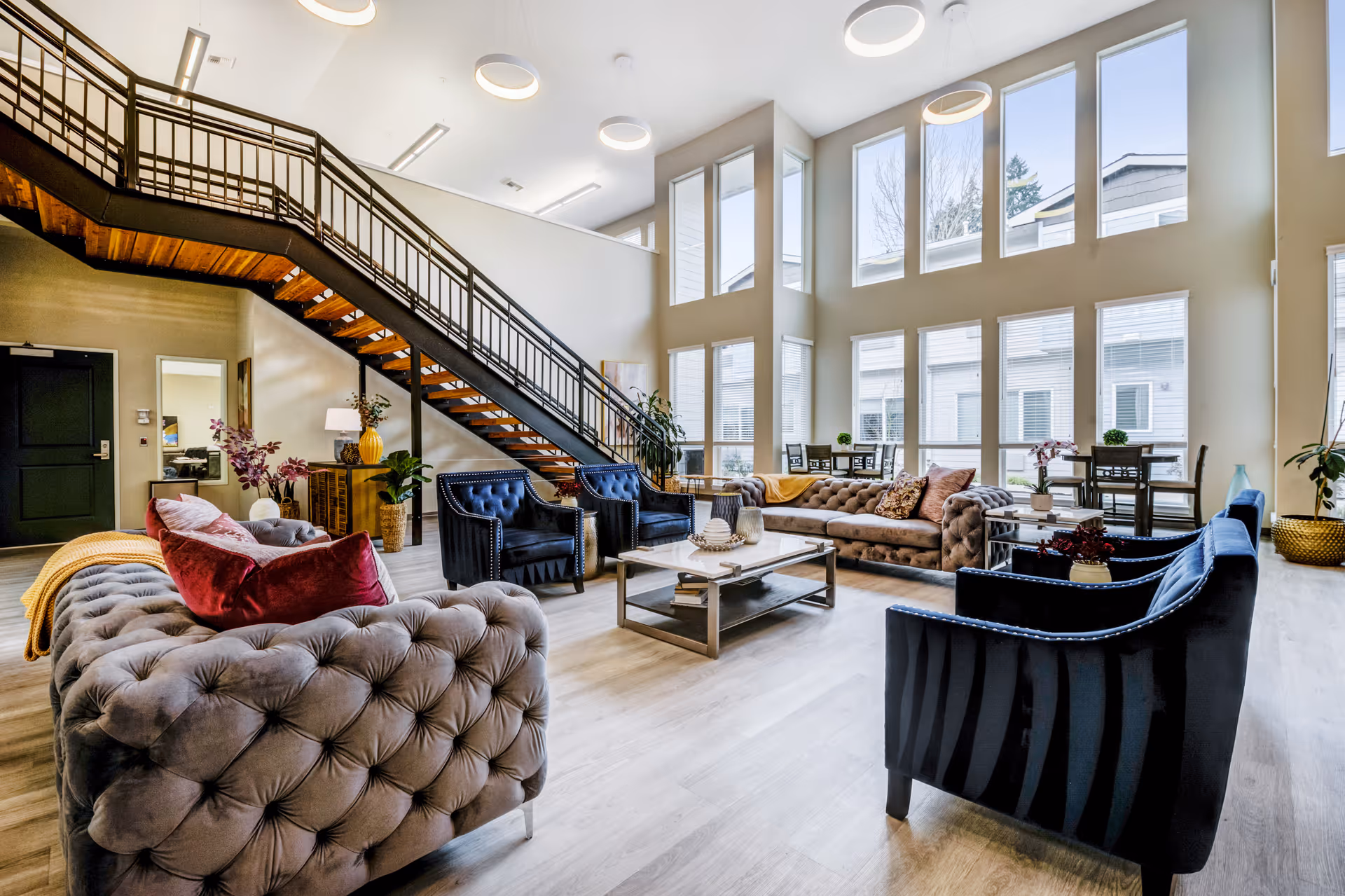 A spacious and well-lit senior living facility common area with high ceilings and large windows. The room features comfortable tufted sofas with colorful pillows, several blue armchairs, a coffee table with decorative items, and a staircase with a wooden underside and metal railing. There are also small dining tables near the windows and various plants and lamps adding to the cozy atmosphere.