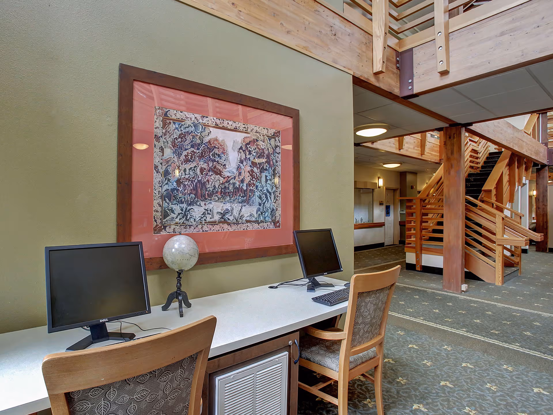 Interior view of a senior living facility showing a workspace with two computer monitors and chairs on a white desk. A decorative globe is placed between the monitors. Behind the desk is a green wall with a large framed artwork depicting a nature scene. To the right, there is a wooden staircase leading to an upper floor, with exposed wooden beams and carpeted flooring with a floral pattern.