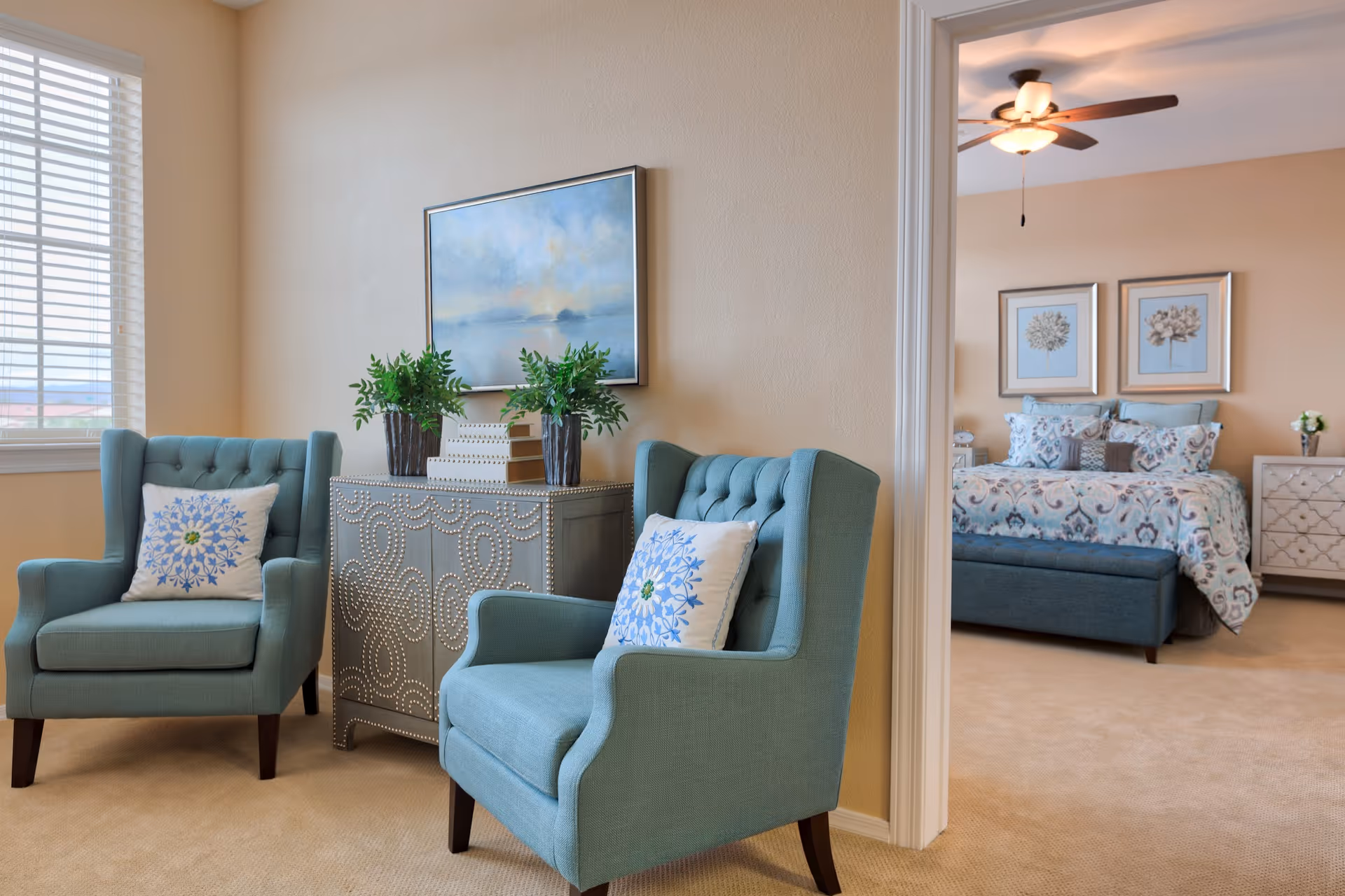 A cozy sitting area with two teal armchairs, each with a decorative pillow featuring a blue floral pattern. Between the chairs is a silver cabinet adorned with two potted plants and a decorative box. A framed landscape painting hangs above the cabinet. Through an open doorway, a bedroom is visible with a bed covered in a patterned comforter, two framed floral artworks on the wall, a ceiling fan, and a nightstand with a small vase of flowers.