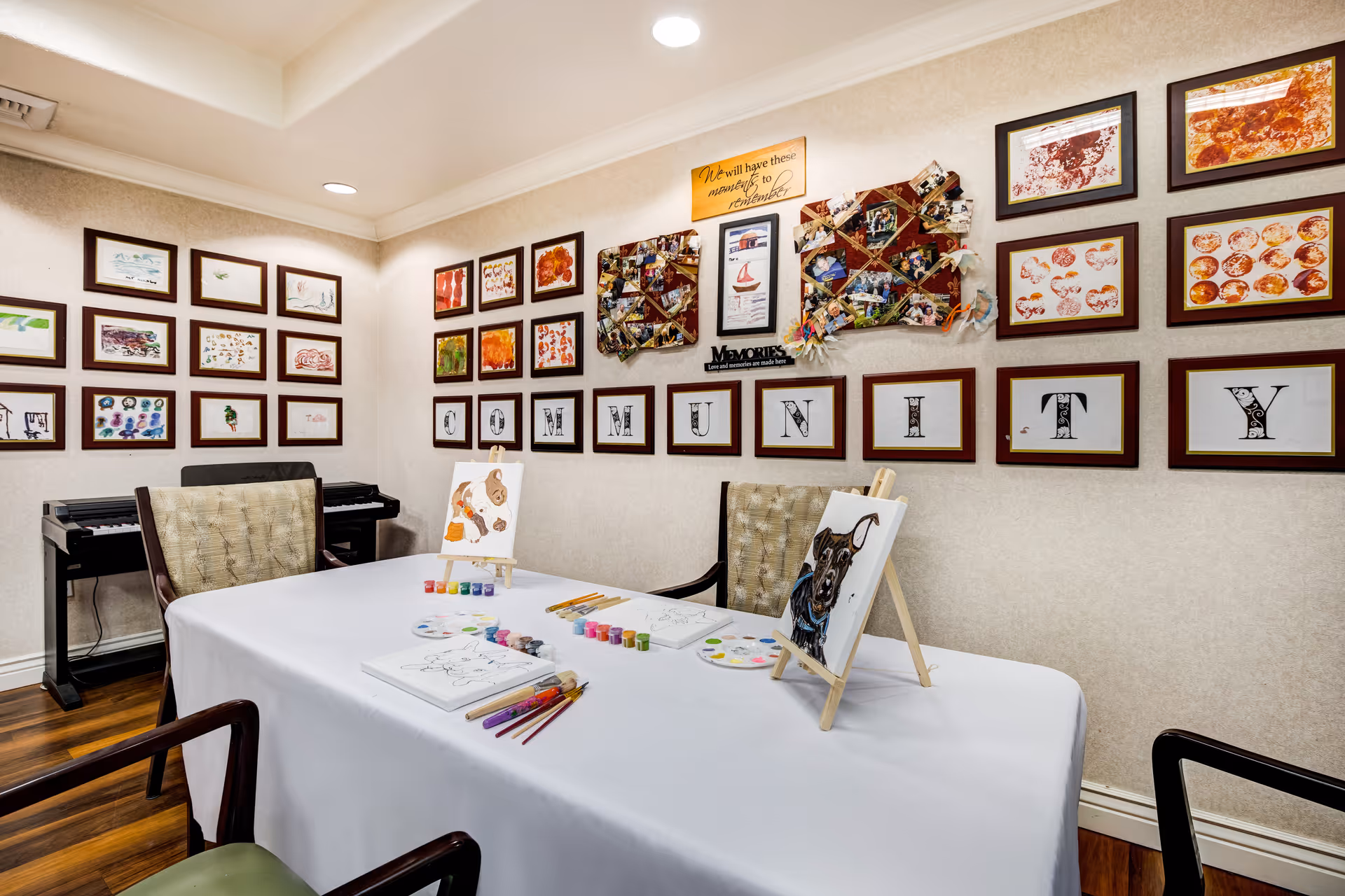 A well-lit room with a table covered by a white cloth, set up for painting with brushes, paint palettes, and canvases featuring dog paintings. The walls are decorated with framed artwork and photos, including a large display spelling out the word 'COMMUNITY'. There is a keyboard against the wall and several chairs around the table.