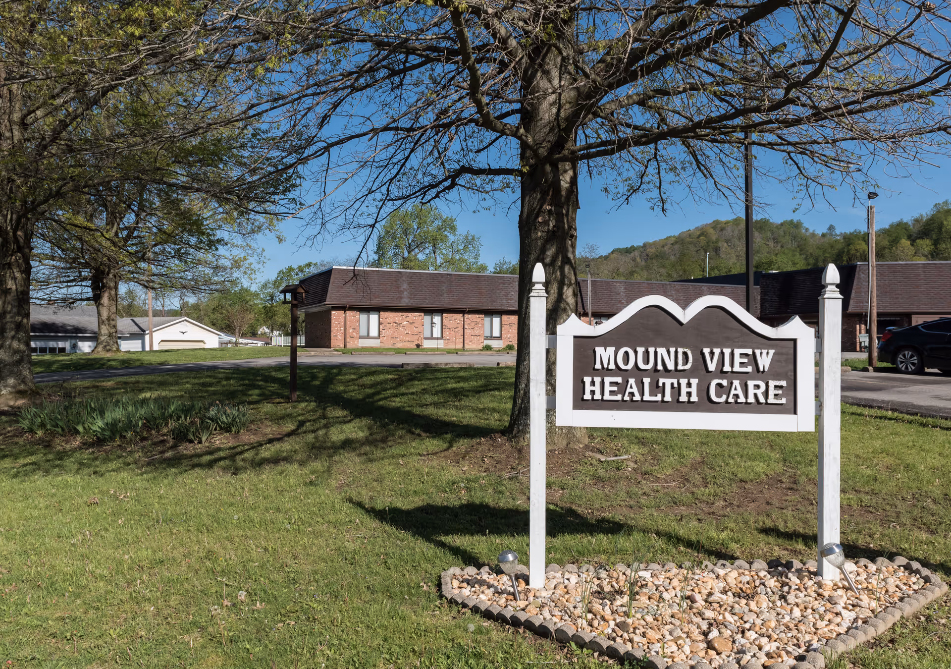 Outdoor view of Mound View Health Care facility with a white and brown sign in the foreground surrounded by small rocks and grass. Trees and a building with a brown roof and brick walls are visible in the background under a clear blue sky.