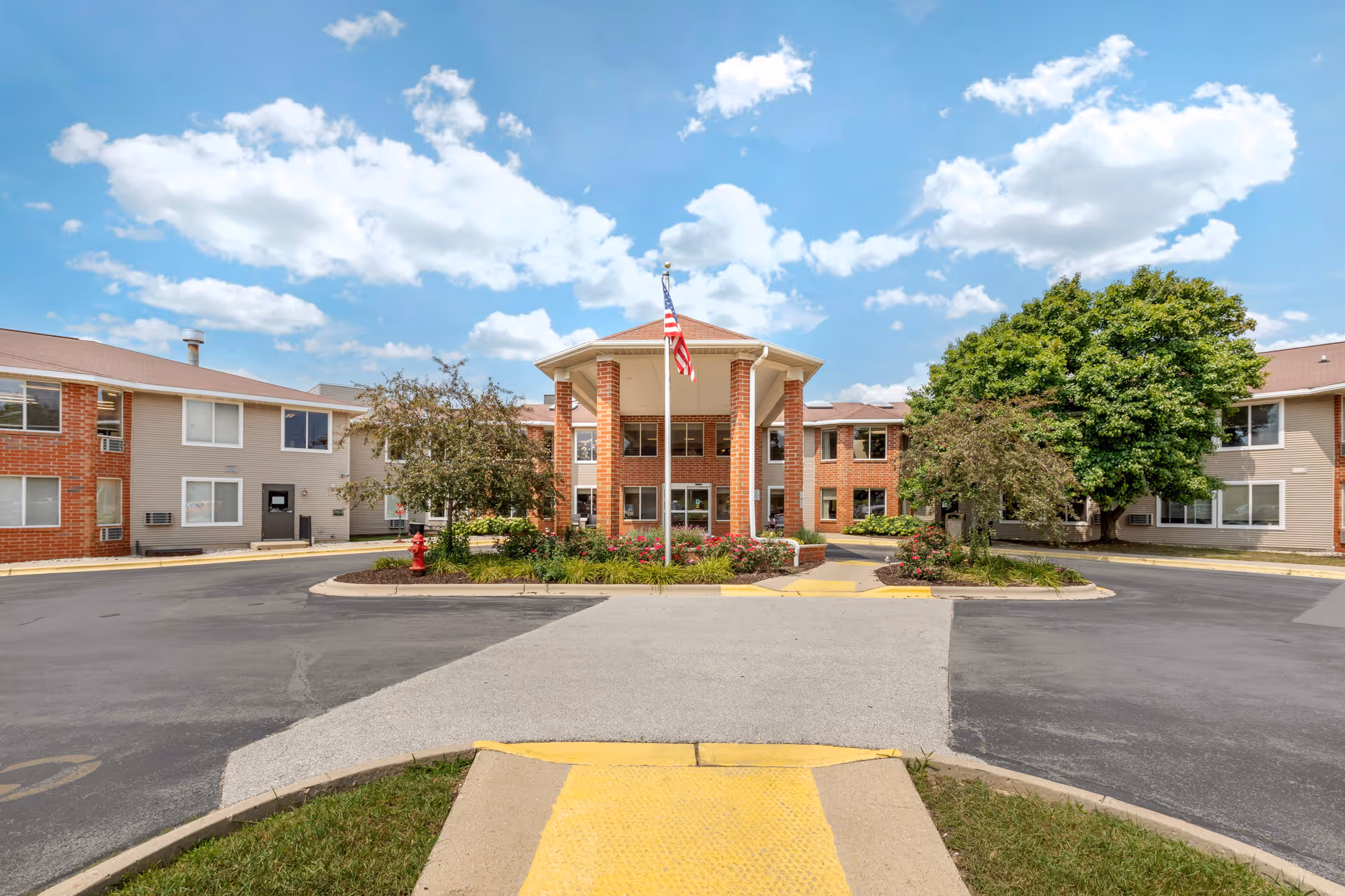 Front exterior view of Layton Terrace Senior Living building with a circular driveway, landscaped garden with flowers and trees, and an American flag on a flagpole under a partly cloudy sky.