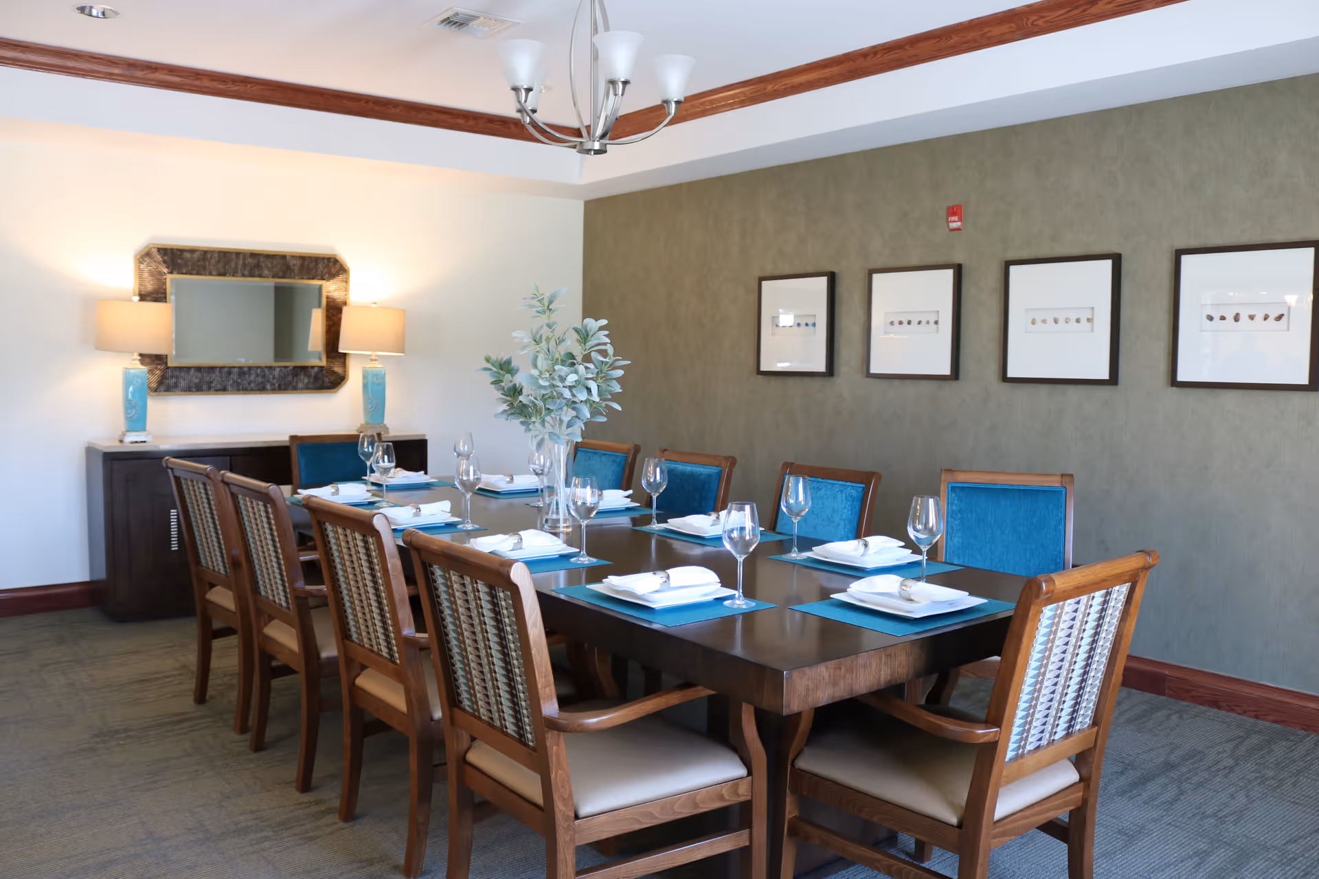 A formal dining room with a long wooden table set for eight people. The table has blue placemats, white plates, folded napkins with napkin rings, and empty wine glasses. There is a centerpiece with green foliage in a clear vase. The room features a green accent wall with four framed artworks, a sideboard with two blue lamps, and a large mirror above it. A modern chandelier hangs from the ceiling.