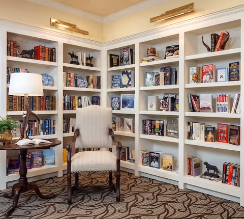 A cozy reading nook featuring a cushioned armchair with wooden arms placed in front of white built-in bookshelves filled with books, decorative plates, and small sculptures. A round wooden side table with a lamp and an open book is positioned next to the chair. The floor is carpeted with a patterned design.