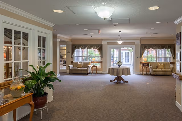 A spacious and well-lit common area in a senior living facility with beige walls and carpeted floor. The room features two striped sofas with pillows, a round table with a floral centerpiece in the center, and large windows with valances letting in natural light. There is a potted plant on a small table to the left and a wooden side table with a glass beverage dispenser. Ceiling lights illuminate the room.