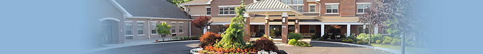 Front entrance of a brick assisted-living building with a circular driveway, landscaped flower beds, and a covered drop-off porte-cochere.