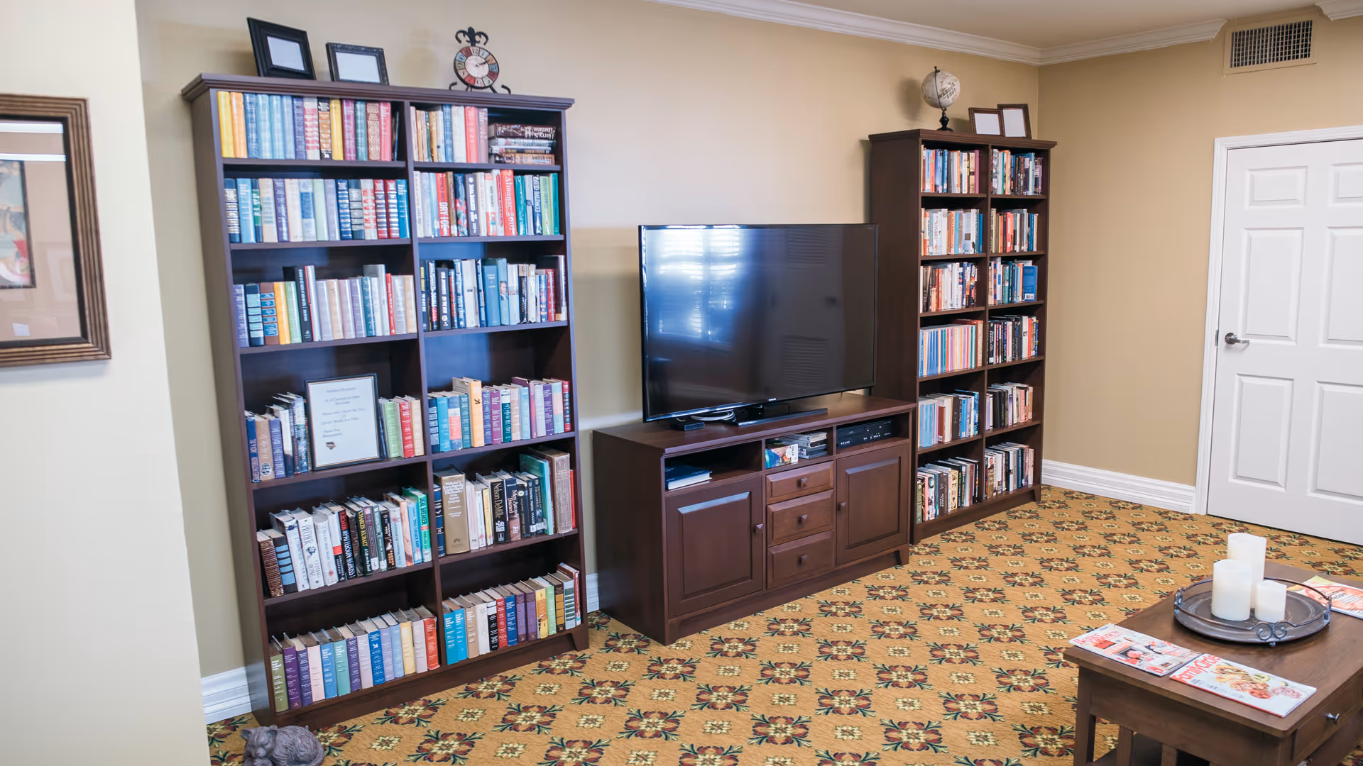 Cozy common room with tall bookshelves, a flat-screen TV on a wooden cabinet, and a coffee table with candles and magazines.