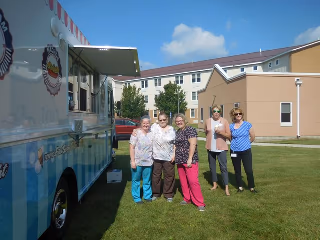Five women standing on a grassy lawn outside a multi-story beige building under a blue sky with some clouds. To the left is a blue and white food truck with a serving window open. The women are casually dressed and smiling, enjoying the outdoor setting.