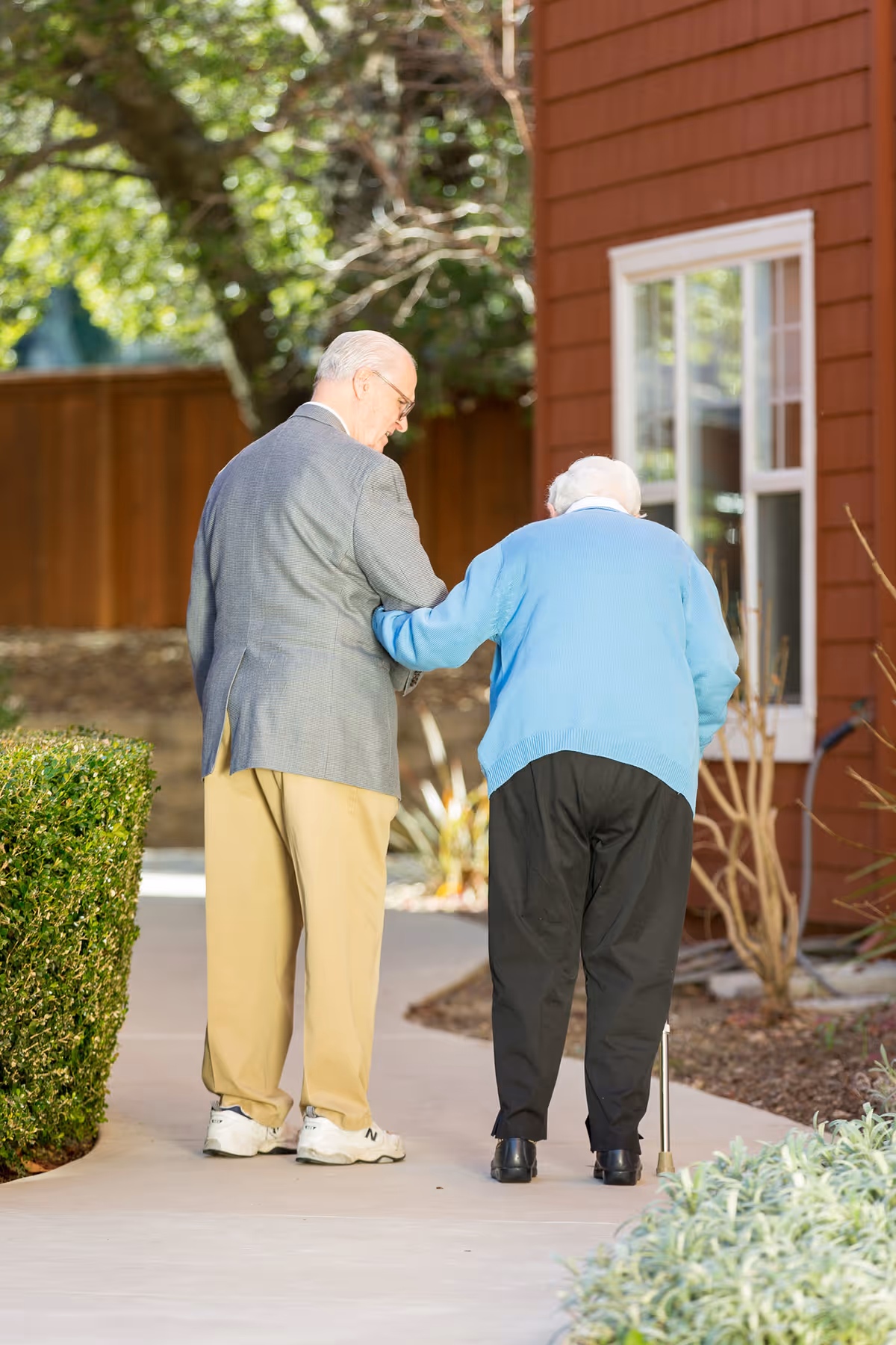 An elderly man in a gray jacket and beige pants is gently supporting an elderly woman wearing a blue sweater and black pants as they walk together on a paved outdoor path near a red building with a window. The woman is using a cane for support.