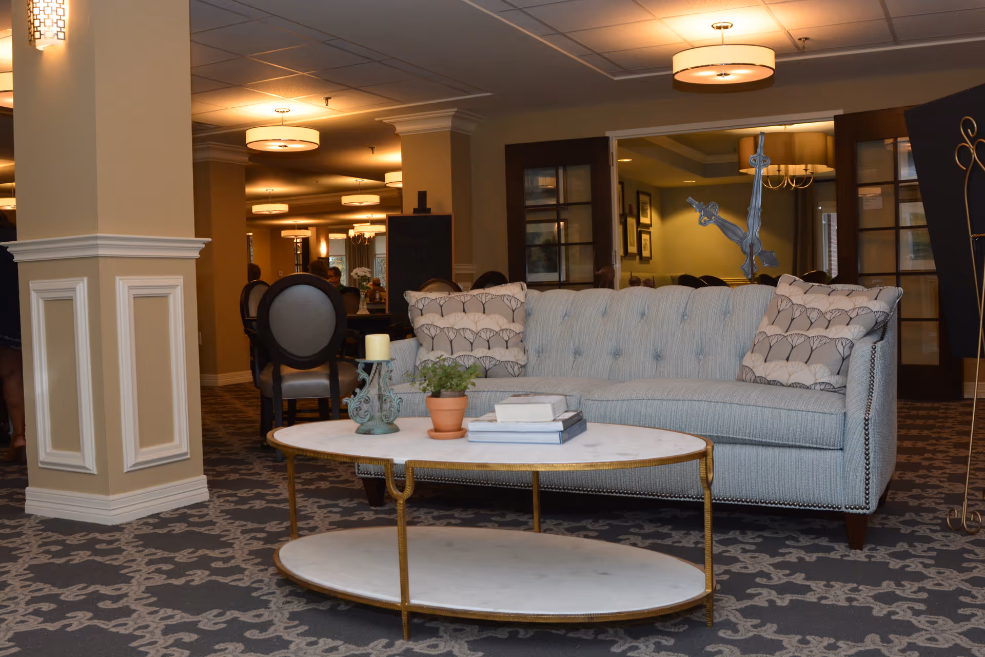 A cozy living room area in a senior living facility featuring a light blue tufted sofa with patterned pillows, a white oval coffee table with gold legs holding a potted plant, books, and a candle holder. The room has beige walls, decorative columns, patterned carpet, and warm ceiling lights. In the background, there are dining chairs and tables, and glass doors leading to another room.