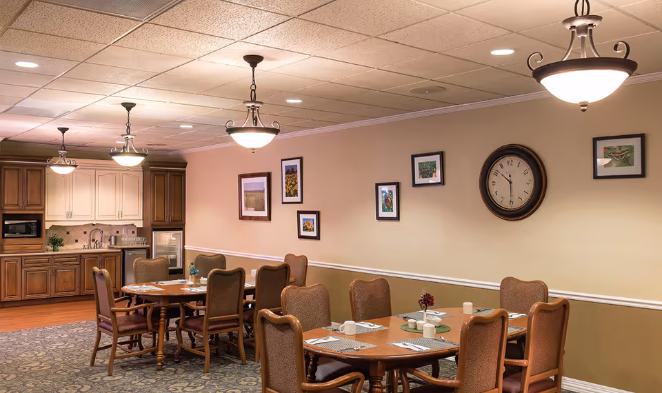 A dining room in Arkansas City Presbyterian Manor featuring two wooden dining tables with chairs arranged around them. The tables are set with placemats, cups, and small flower vases. The room has beige walls decorated with framed pictures and a large round clock. There are three hanging light fixtures on the ceiling, and a kitchenette area with wooden cabinets, a microwave, a sink, and a small refrigerator is visible in the background.