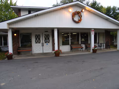 Front exterior view of a single-story building with white siding and brick accents. The entrance has double doors with diamond-shaped window panes, flanked by benches and potted plants with flowers. A decorative wreath is hung above the entrance under the roof peak. There are outdoor lights on either side of the entrance and a no parking sign on a column to the right.
