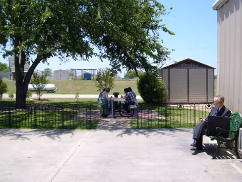 Outdoor area with a concrete patio and green grass. Two people are sitting at a picnic table under a tree, and another person is sitting alone on a green bench near a building. There is a small shed and some fencing in the background under a clear blue sky.