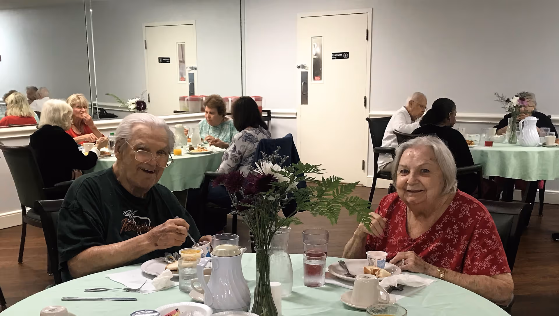 Elderly residents sitting at round tables covered with light green tablecloths, eating and socializing in a dining room. The room has white walls, a large mirror, and a door labeled 'Employees Only'. There are flowers in a vase on the table in the foreground.