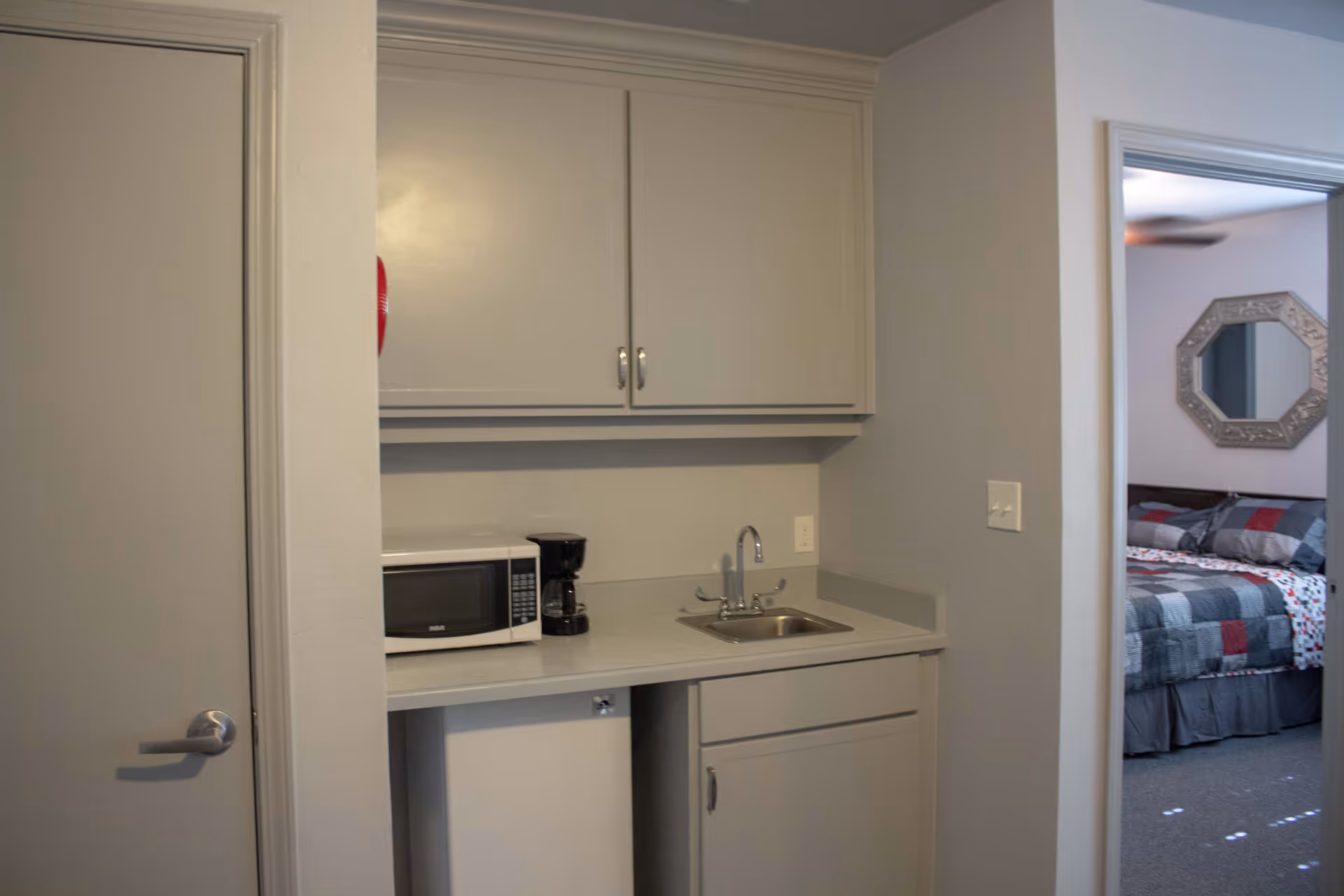 Small kitchenette with microwave, coffee maker, sink and cabinets next to a doorway showing a bedroom.