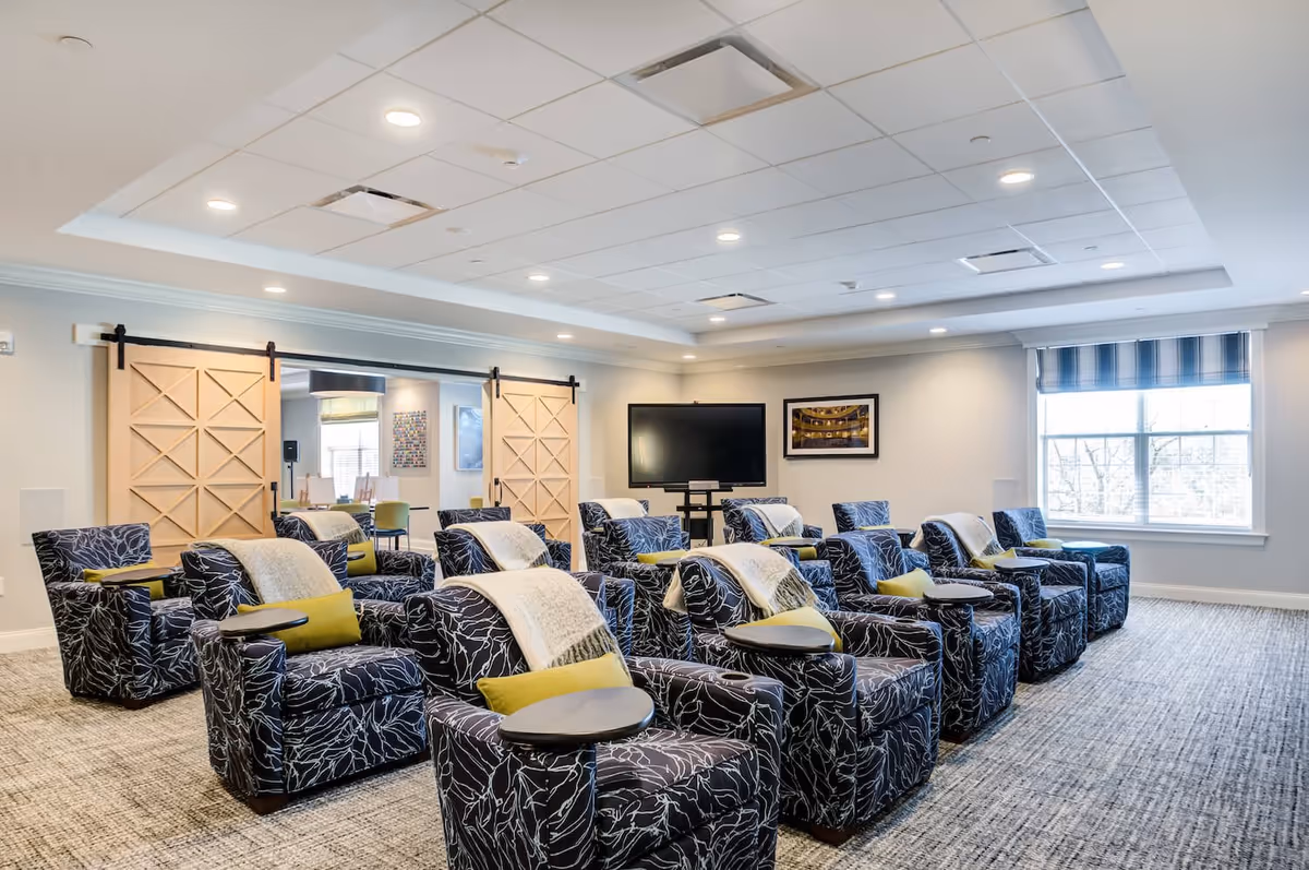 A bright and spacious common area with multiple patterned armchairs arranged in rows facing a large flat-screen TV mounted on a stand. Each chair has a small attached table, a green cushion, and a light-colored throw blanket. The room features a large window with striped blinds, recessed ceiling lights, and sliding barn doors leading to another room with additional seating and artwork on the walls.