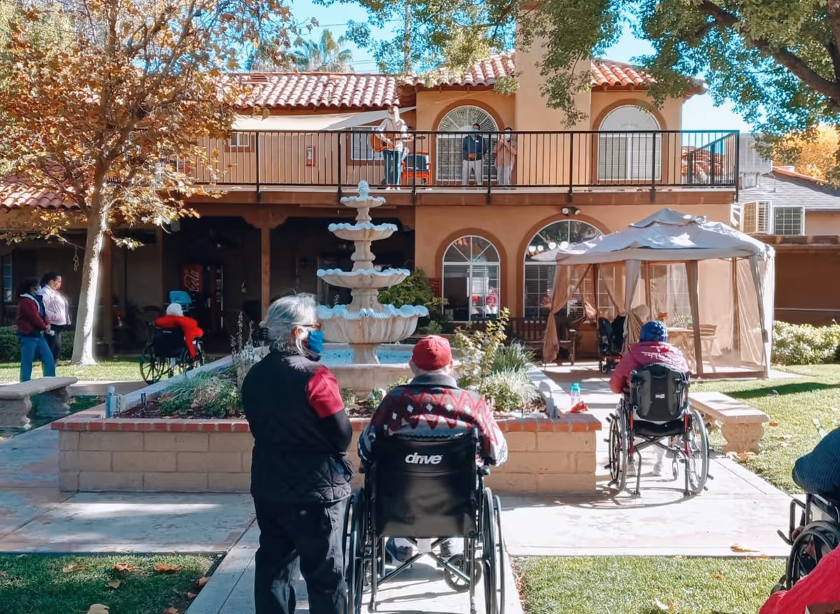 Outdoor courtyard area of a senior living facility with a central multi-tiered fountain, several seniors in wheelchairs and standing, some wearing masks, a gazebo with seating, and a two-story building with a balcony in the background.