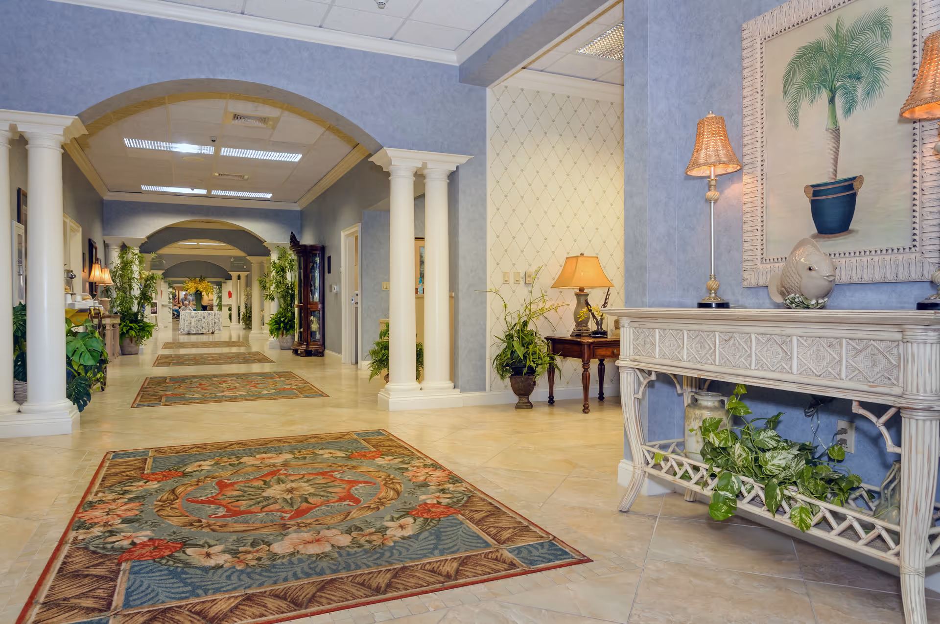 Bright, elegant interior hallway with decorative columns, patterned rugs, plants, and console tables.