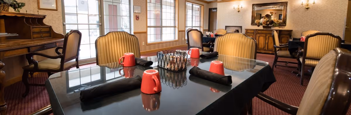 A dining room with a glass-topped table set with red mugs and black napkins and upholstered chairs around it.