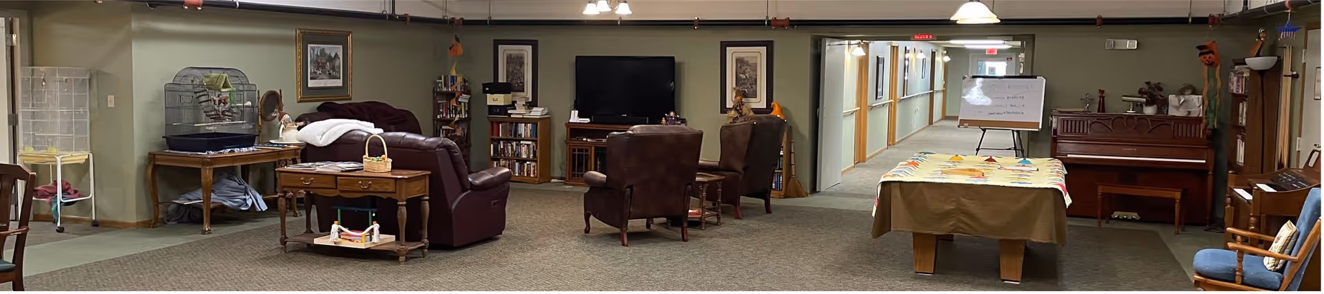 A spacious assisted living common area with comfortable seating including leather armchairs and a sofa, a television mounted on the wall, bookshelves filled with books, a piano, and a table covered with a colorful cloth. The room has green walls and carpeted floors, with a hallway leading to other rooms in the background.