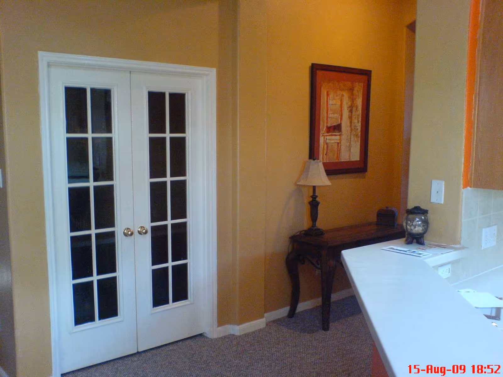 Interior view of a room with beige walls featuring white double French doors with glass panes, a small wooden table with a lamp and decorative items, and a white countertop in the foreground.