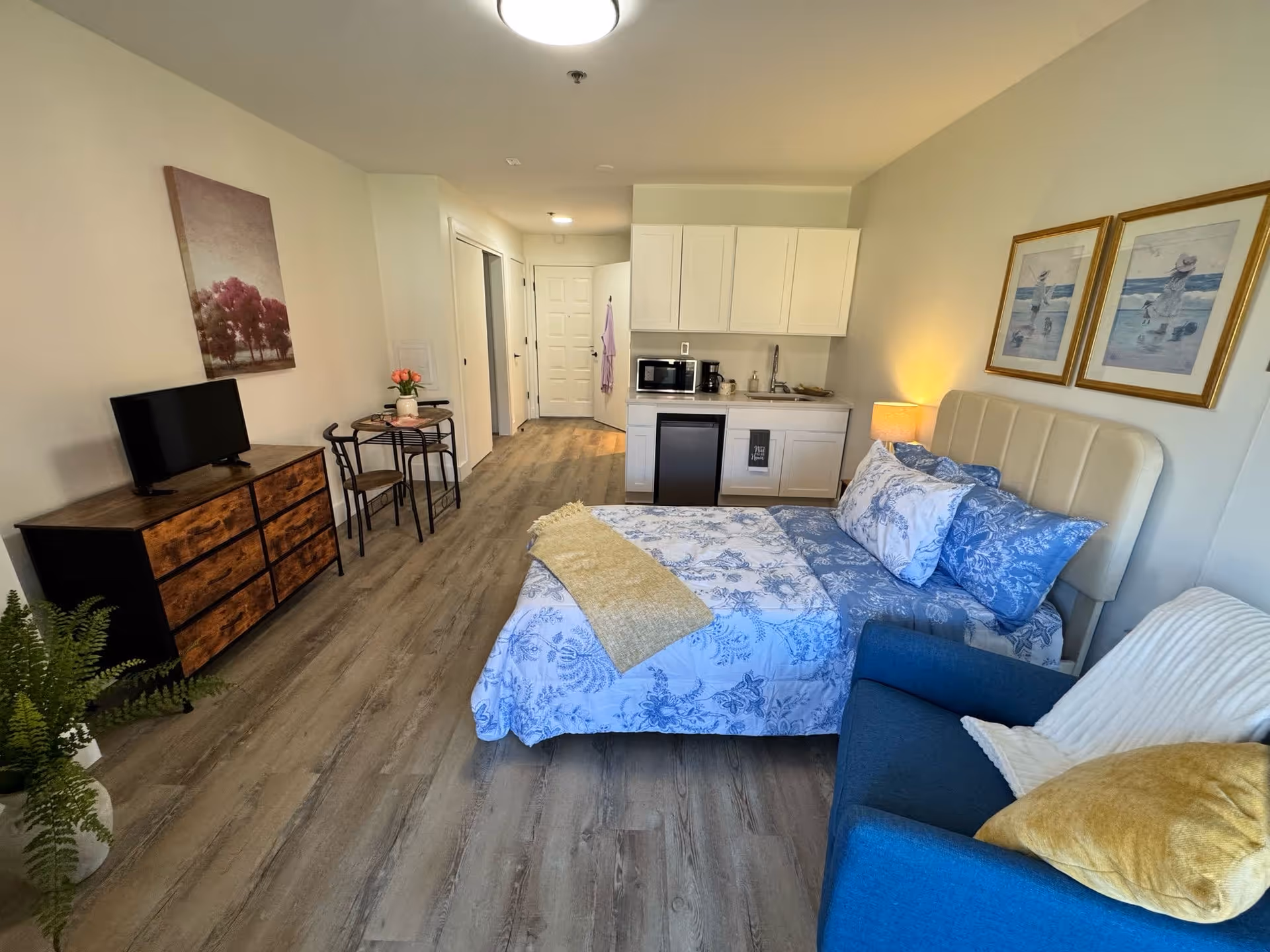 A studio apartment interior at The Fountains at Atco featuring a bed with blue and white floral bedding, a blue armchair with yellow and white pillows, a small kitchenette with white cabinets, a microwave, and a mini fridge. There is a wooden dresser with a TV on top, a small round dining table with two chairs, and framed artwork on the walls. The floor is wood laminate and the room is well-lit with ceiling lights.