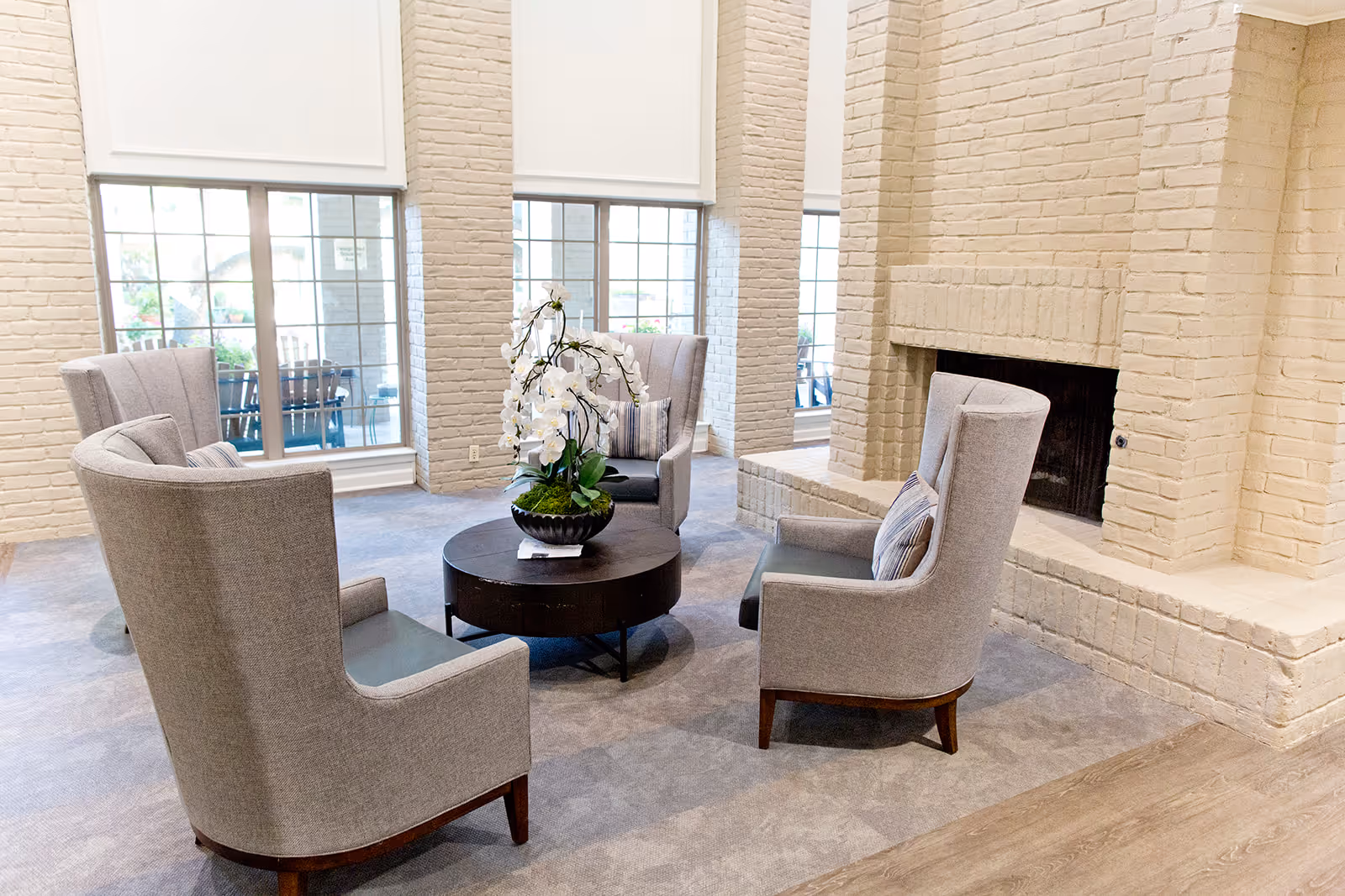 A bright and cozy sitting area with four gray upholstered armchairs arranged around a round dark wooden coffee table with a white orchid centerpiece. The room features large windows with white blinds, white brick walls, and a white brick fireplace.