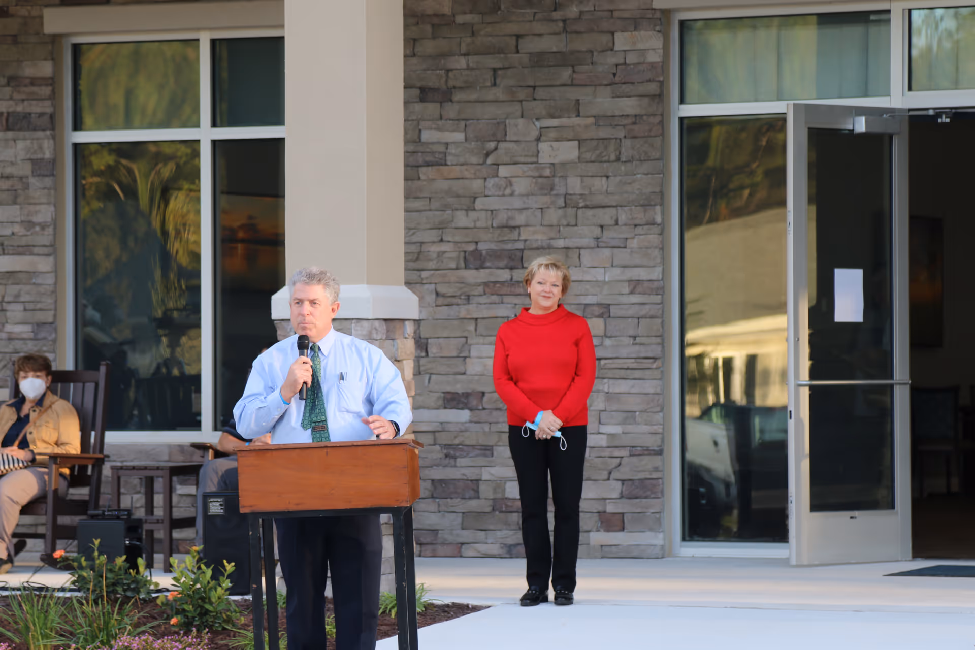 A man speaks at a podium in front of a stone-faced building entrance while a woman stands behind him and others sit nearby.