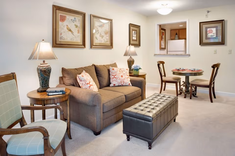 A cozy living room area with a brown loveseat adorned with patterned pillows, a wooden side table with a decorative lamp and books, a cushioned armchair, and a tufted leather ottoman. In the background, there is a small round dining table set for two with wooden chairs, and a pass-through window to a kitchen with a refrigerator. The walls are decorated with framed artwork featuring birds and flowers.