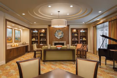 A spacious and elegantly decorated living room area with a round ceiling light fixture, built-in wooden shelves with decorative items, a fireplace, a green sofa, several chairs, a grand piano near large windows with striped curtains, and a patterned carpet.