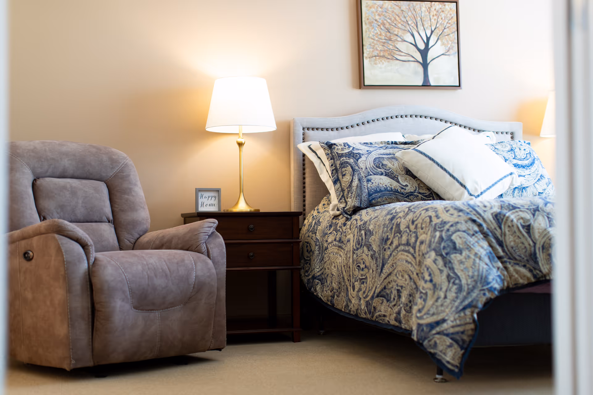 A cozy bedroom corner with a paisley-patterned bed, a wooden nightstand and lamp, an upholstered recliner, and framed tree artwork on the wall.