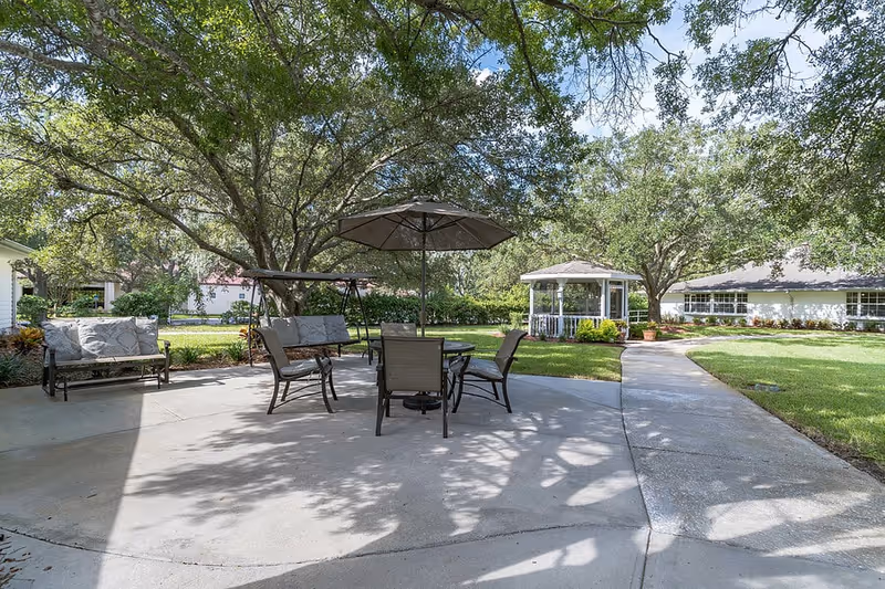 Outdoor patio area with cushioned benches, chairs, a table with an umbrella, and a swing under large trees. A paved walkway leads to a white gazebo surrounded by green grass and trees.