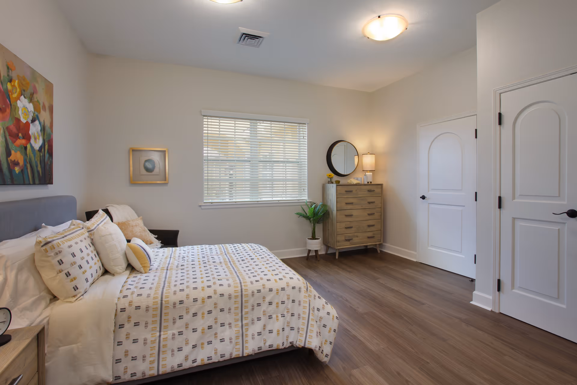 A bright and clean bedroom with a bed featuring patterned bedding and multiple pillows. There is a wooden dresser with a round mirror and a lamp on top, a potted plant beside it, two white doors on the right wall, and a window with blinds letting in natural light. The walls are painted light beige, and there is a colorful floral painting above the bed.