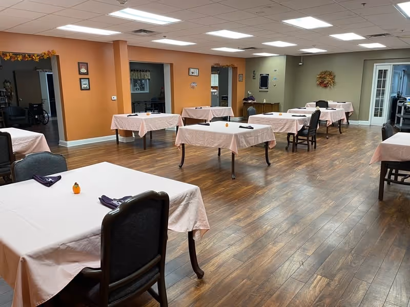 A dining room in a senior living community with several tables covered in light pink tablecloths, each set with a small decorative pumpkin and folded dark napkins. The room has wood flooring, orange and beige walls, and multiple chairs around the tables. There are doorways and wall decorations visible in the background.