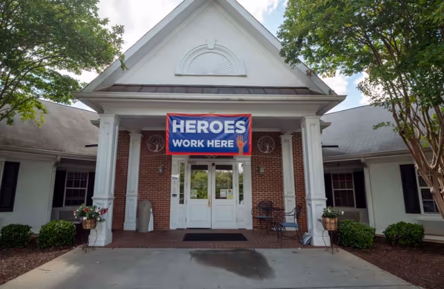 Front entrance of a brick and white building with a covered porch supported by white columns. A large banner hanging above the double glass doors reads 'HEROES WORK HERE' with a red hand graphic. There are potted plants on either side of the entrance and trees framing the building.