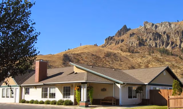Single-story senior living facility building with beige siding and a brown roof, featuring a covered entrance with a bench and some greenery. In the background, there is a rocky, tree-covered hillside under a clear blue sky.