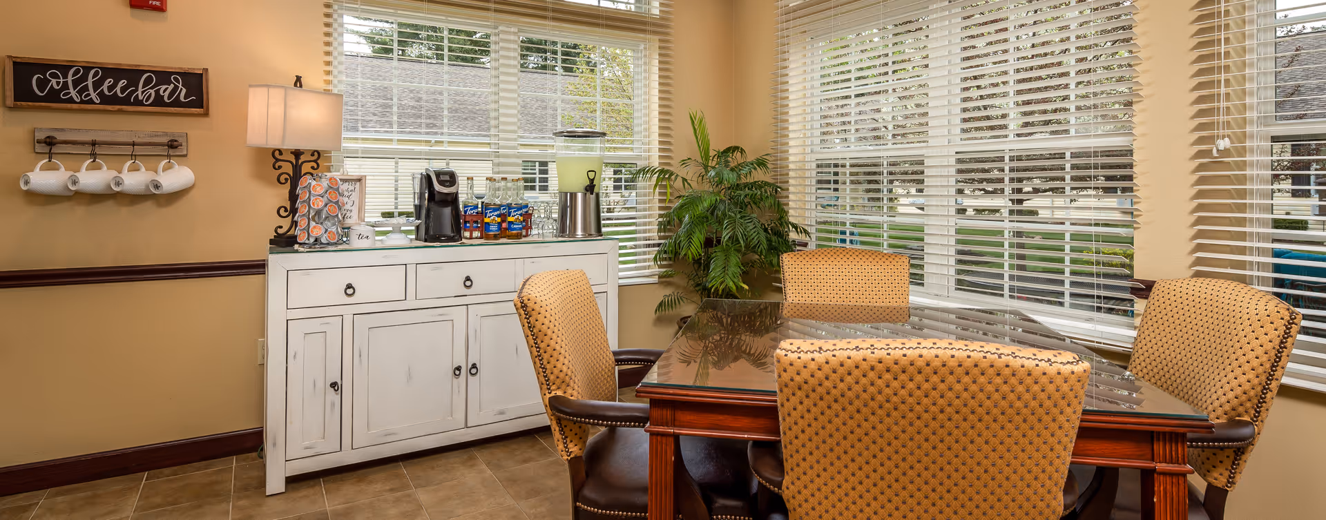 A cozy dining area with a wooden table and four upholstered chairs. There is a white cabinet against the wall with a coffee maker, cups, and other beverage supplies on top. A sign above the cabinet reads 'coffee bar'. Large windows with blinds allow natural light to fill the room, and a green plant is placed in the corner.