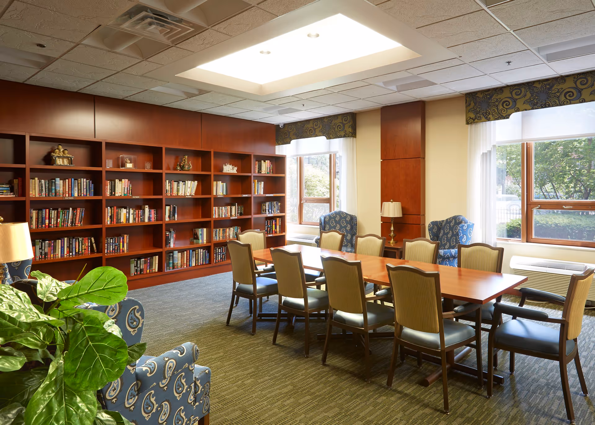 A well-lit room with a long wooden table surrounded by chairs, two blue patterned armchairs near the windows, a large wooden bookshelf filled with books along one wall, and a green leafy plant in the foreground. The room has large windows with patterned valances and a carpeted floor.