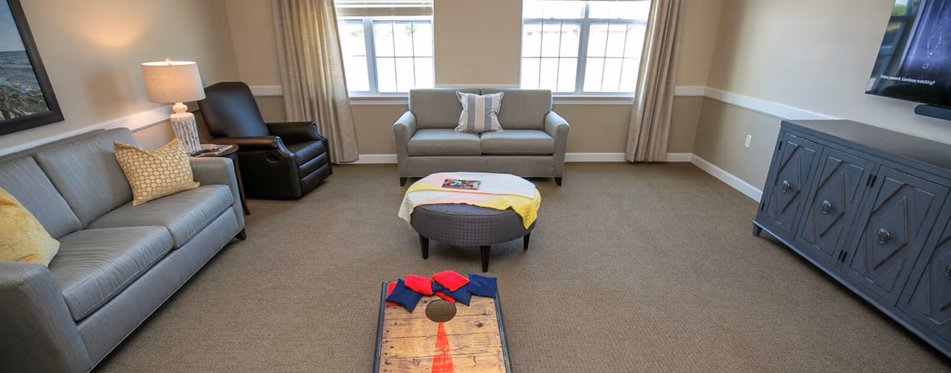 Bright common living room with sofas, an armchair, an ottoman, a TV cabinet, and a cornhole game on the carpet.
