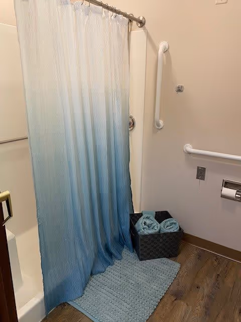 Bathroom with a white and blue ombre shower curtain, a light blue bath mat on a wooden floor, a black basket containing rolled light blue towels, and white grab bars on the walls.