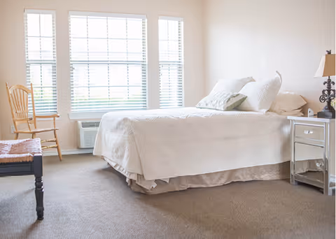 A bright bedroom with a large bed covered in white bedding and pillows, a wooden chair near the window, a small bedside table with a lamp, and carpeted floor. Three large windows with blinds allow natural light to fill the room.