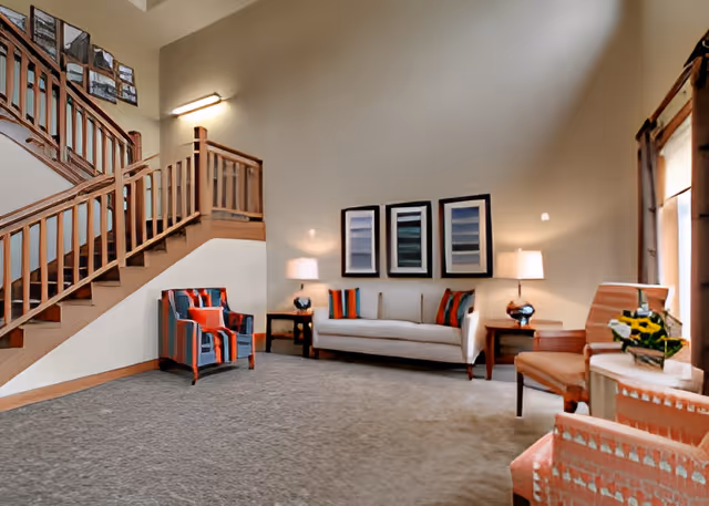 A cozy living room area with a beige sofa adorned with colorful pillows, two matching side tables with lamps, a striped armchair, and two coral-patterned chairs near a window with curtains. A wooden staircase with framed pictures on the wall is visible on the left side.