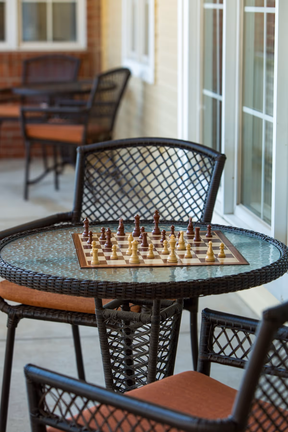 Outdoor patio area with a glass-top round table and wicker chairs with orange cushions. A chessboard with chess pieces set up is placed on the table. The background shows a brick wall and windows of the building.