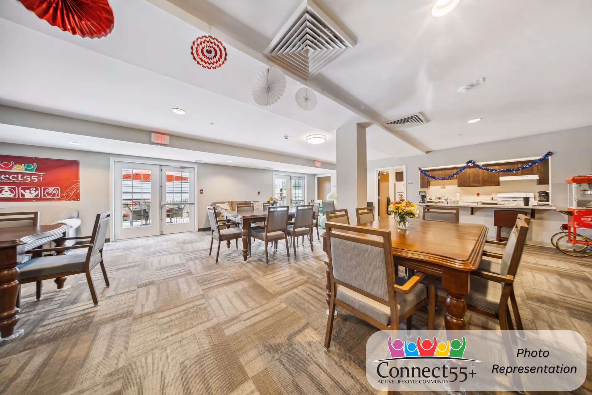 Interior view of a senior living community common area with multiple wooden tables and chairs arranged for dining or socializing. The room has carpeted flooring, ceiling decorations in red and white, and a kitchen area in the background decorated with garlands. There are glass doors leading to an outdoor patio with red umbrellas.
