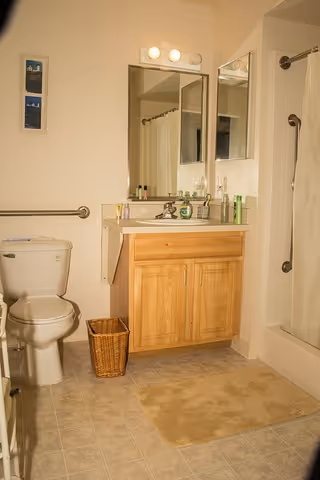 A bathroom with a toilet, wooden vanity with sink and mirror, shower with curtain, and a small wicker wastebasket.