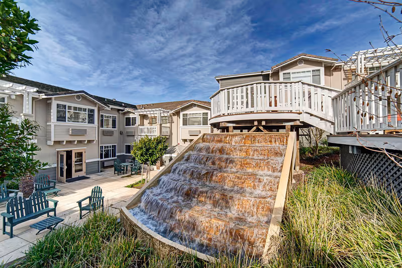 Outdoor courtyard area of Fremont Hills facility featuring a cascading water feature with wooden steps, surrounded by greenery and patio seating with multiple chairs and tables. The background shows a two-story building with balconies and large windows under a partly cloudy blue sky.