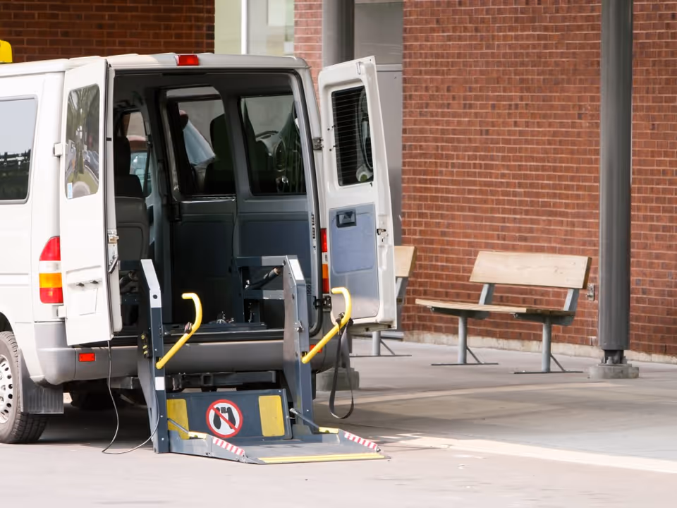 A white van with its rear doors open, showing a wheelchair lift extended to the ground. The van is parked outside a building with a brick wall and two wooden benches nearby.