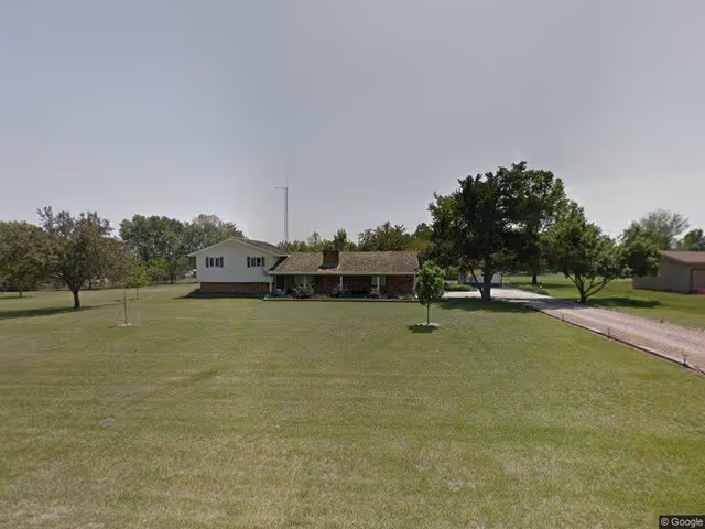 Front view of a single-story ranch house with a large grassy lawn, trees, and a driveway.