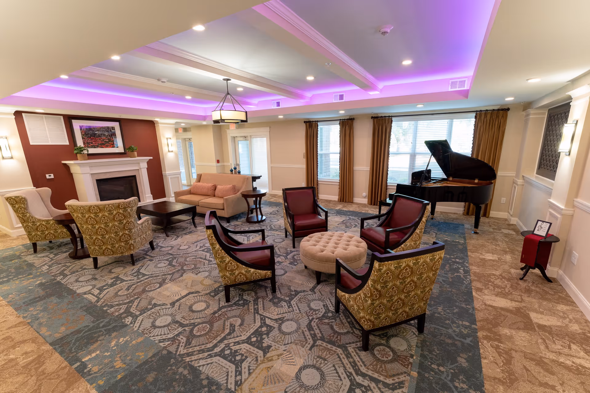 A spacious and well-lit common area in a senior living facility featuring a patterned carpet, multiple upholstered chairs arranged around a round ottoman, a beige sofa, a fireplace with a framed picture above it, large windows with brown curtains, and a black grand piano in the corner. The ceiling has recessed lighting and purple accent lights.